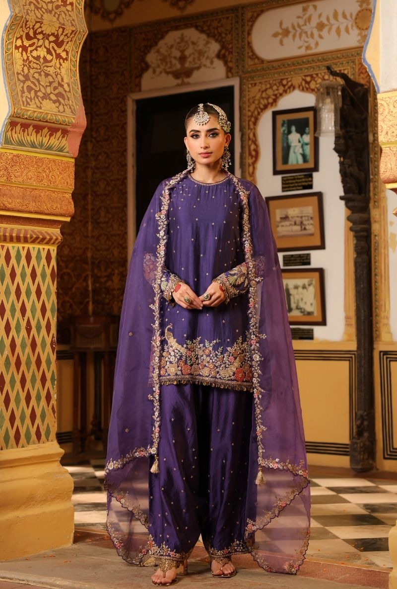 Woman in a traditional purple outfit with intricate designs standing in an ornate interior setting.