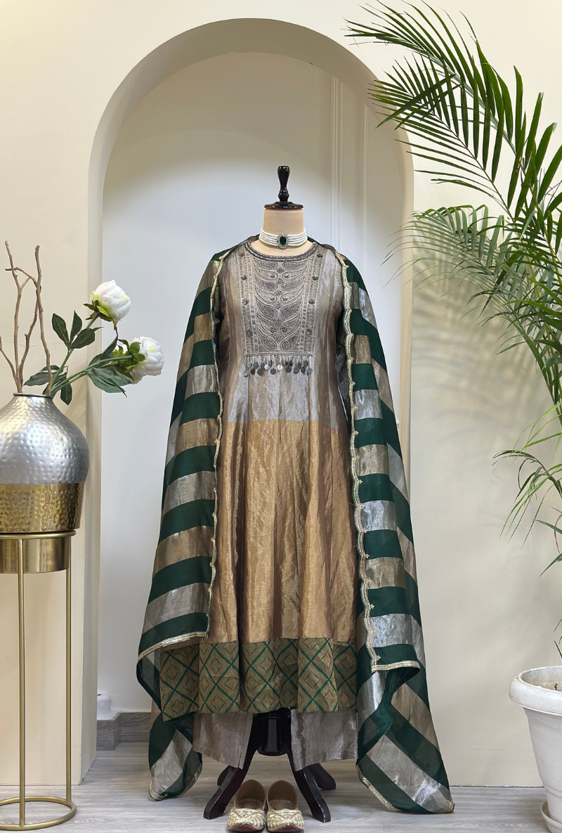 A mannequin displaying a brown Anarkali set with intricate hand embroidery on the front yoke, a plain bottom with an organza patch, and a multi-striped dupatta with sequin detailing.