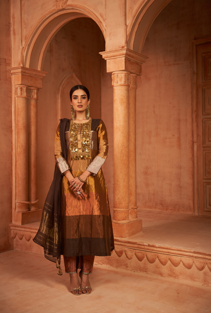 A woman standing in a beautifully decorated arched hallway wearing a traditional brown anarkali set with metallic coin work and a dupatta.