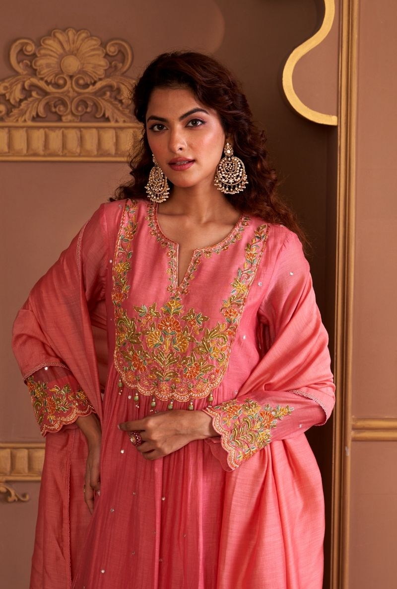 Woman wearing a pink embroidered traditional outfit with gold jewelry against a decorative wall.