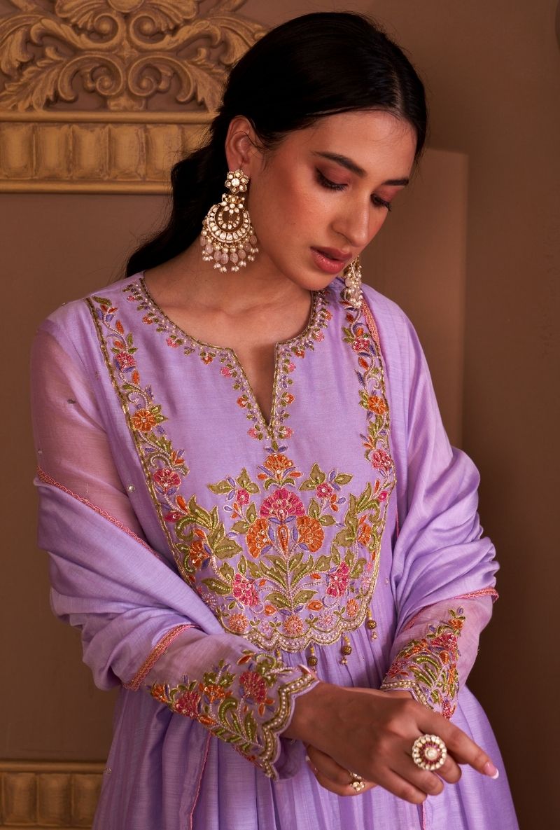 Woman wearing a lavender embroidered outfit with gold jewelry against a beige wall.