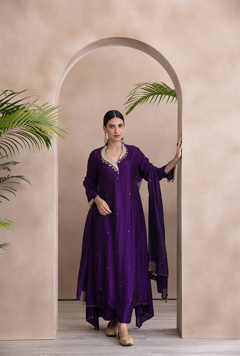 A woman wearing a purple A-line kurta set with leaf buta in zardozi and tikki detail, paired with coordinated lowers. A dupatta is draped over the shoulder.