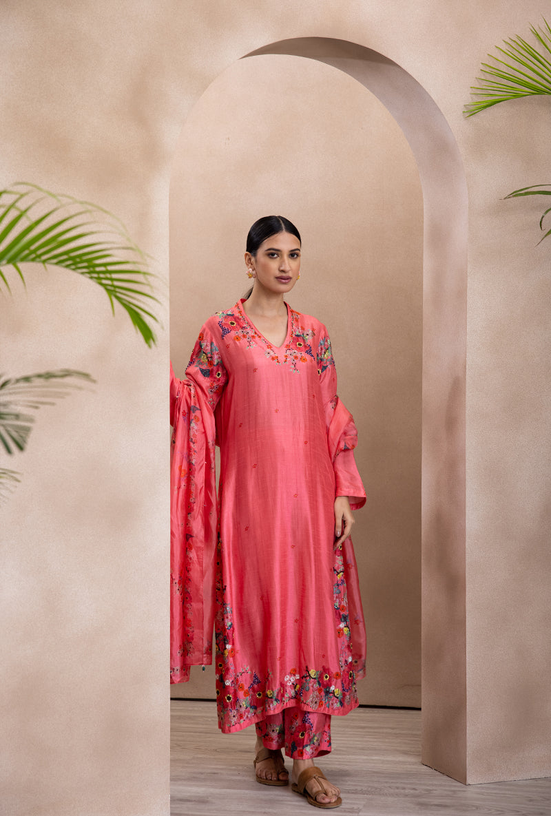 A woman standing in an arched doorway wearing a coral pink V-neck printed bahaar adhira kurta set with floral details and sequins.