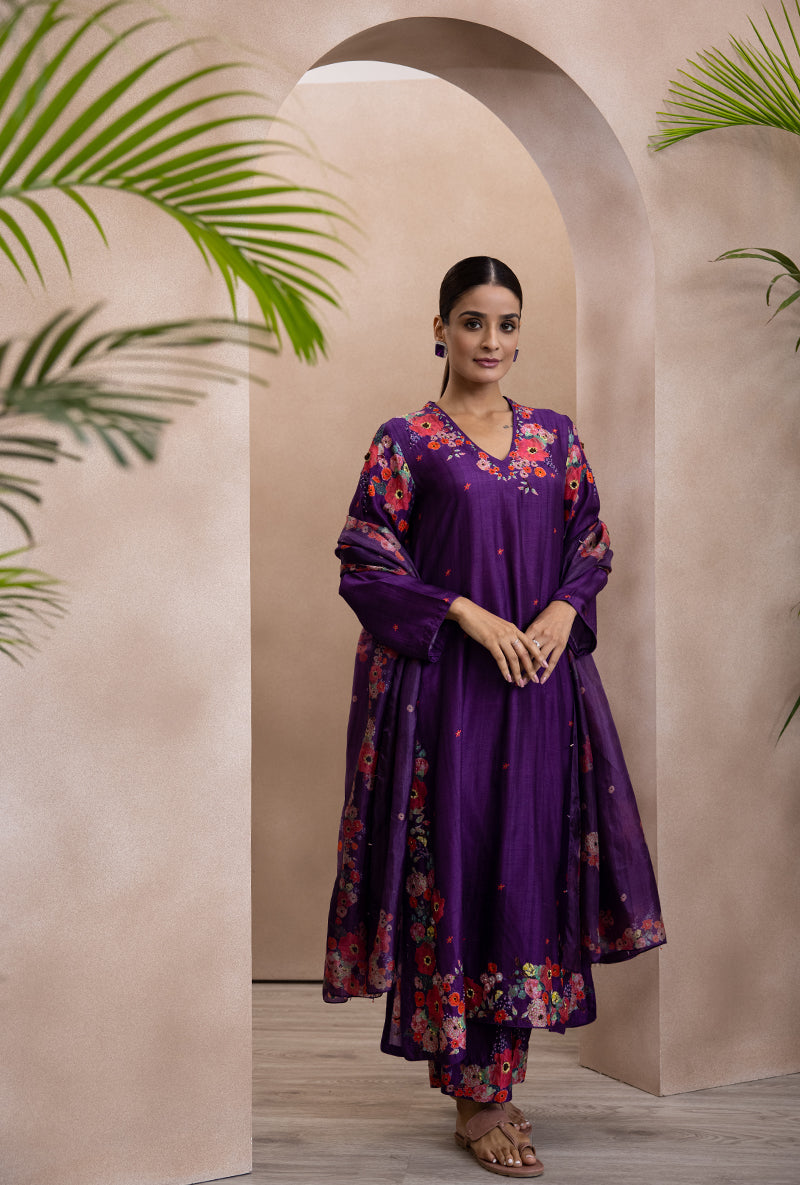 A woman standing in an arched doorway wearing a purple V-neck printed bahaar adhira kurta set with floral patterns and sequins detailing.