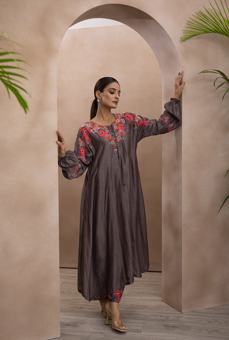 A woman modeling a charcoal grey tie-up printed bahaar adhira kurta set with floral patterns, featuring French knots, sequins, and beads. The set is presented against a backdrop with neutral tones and green foliage.