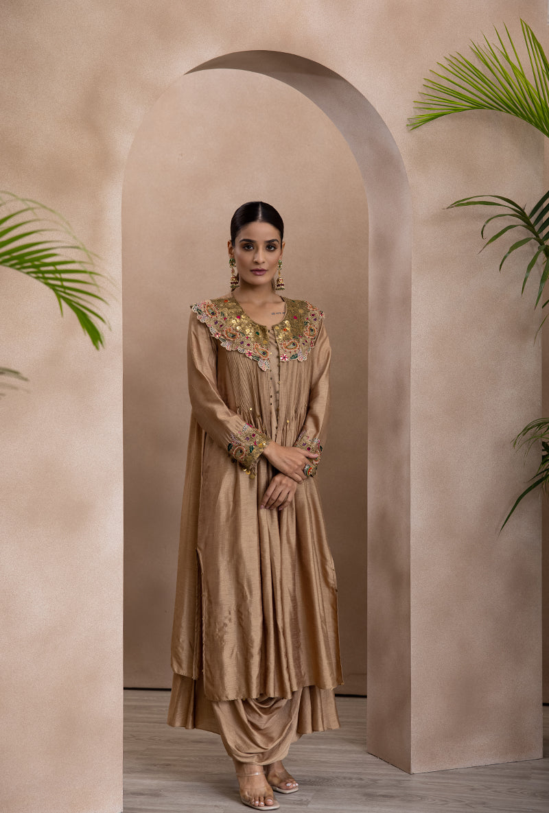 A woman standing in an elegant pose wearing a beige off-shoulder cowl neck jacket and matching skirt with floral embroidery and gota flowers.