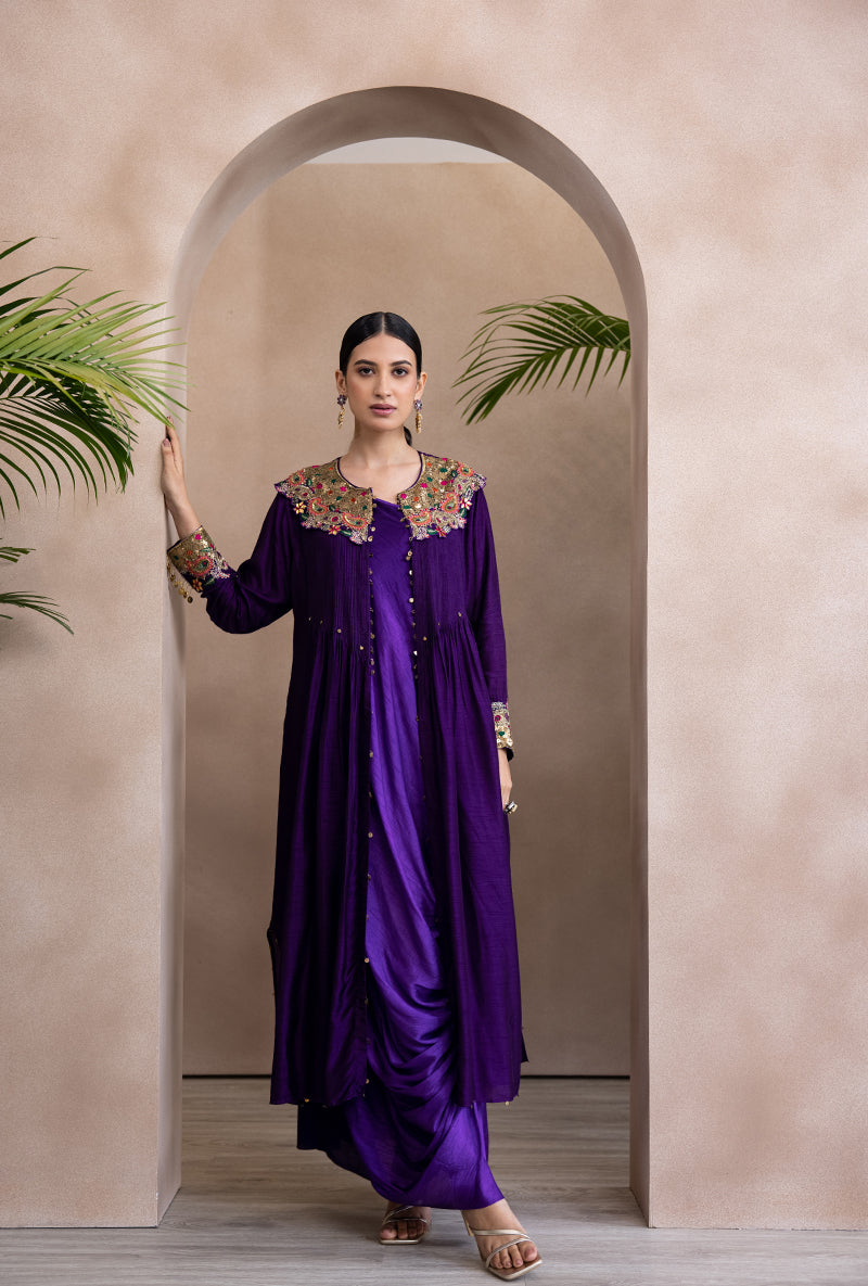 A woman standing in an elegant pose wearing a purple paisley cowl adhira jacket set with embroidery and tassel detailing.