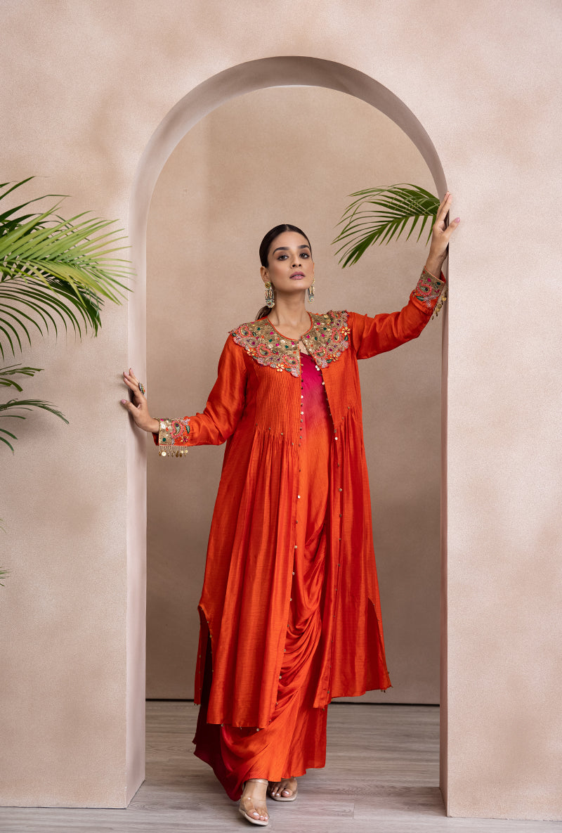 A woman standing in an elegant pose wearing an orange and pink ombre off-shoulder dress with paisley patterns and embellishments, paired with a matching cowl neck jacket.