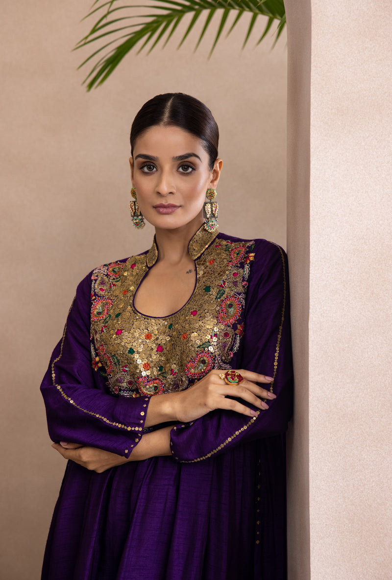 A woman wearing a purple kurta set with detailed embroidery on the yoke, paired with matching lower pants. She is posing indoors with a smile.