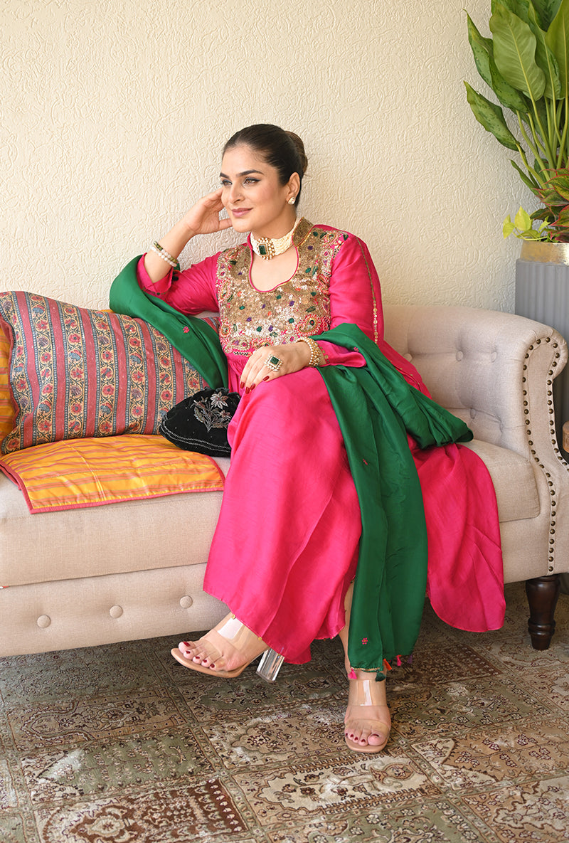 A woman sitting on a couch wearing a pink kurta set with green embellishments and a dupatta.