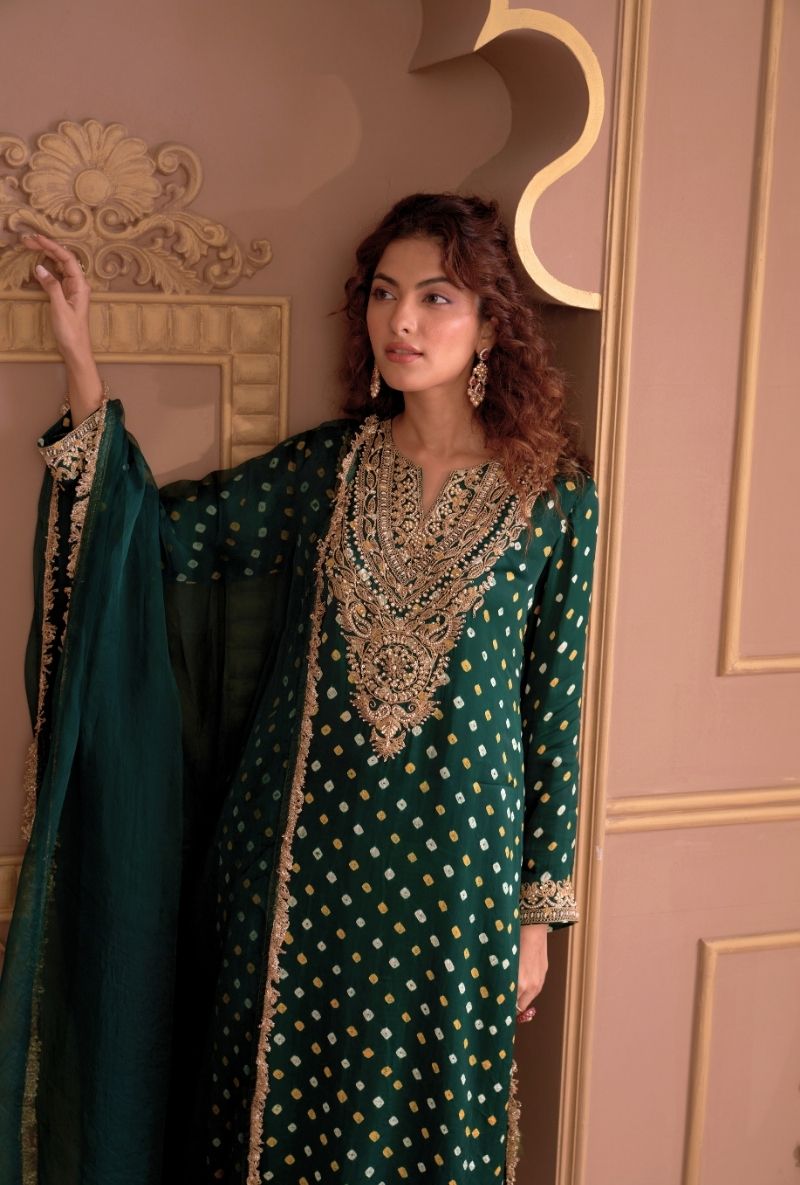 Woman in a green embroidered traditional outfit standing in a decorated room.
