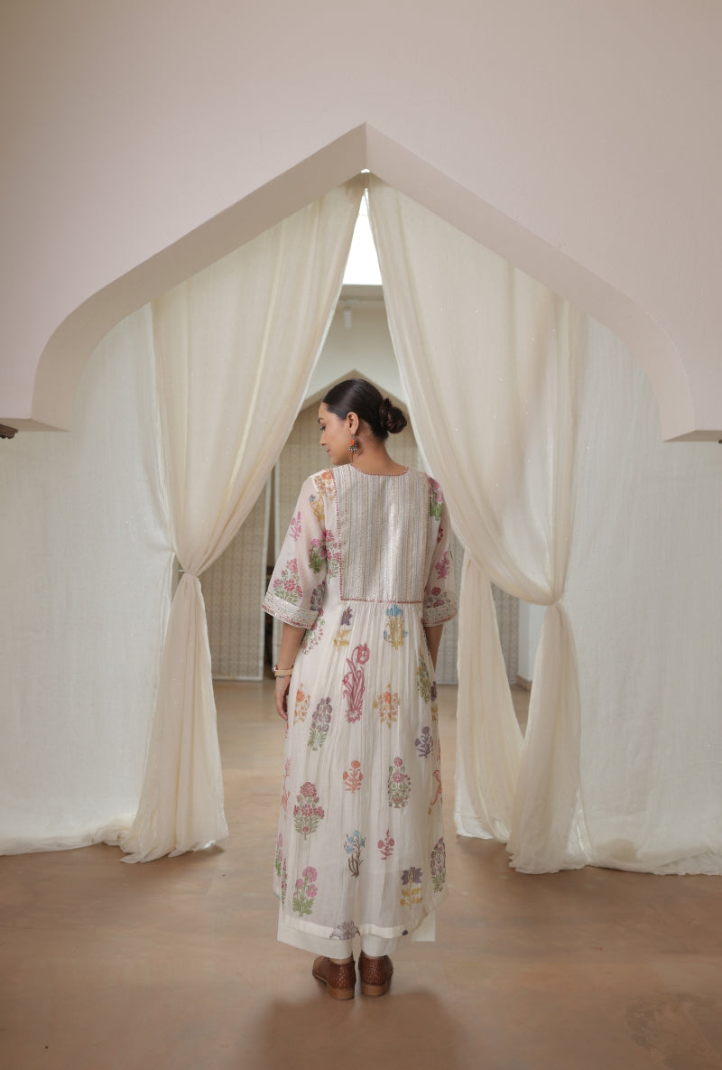 A woman stands in profile wearing a long white kurta dress with a pastel floral print, set against a minimalist interior with draped cream fabric arches.