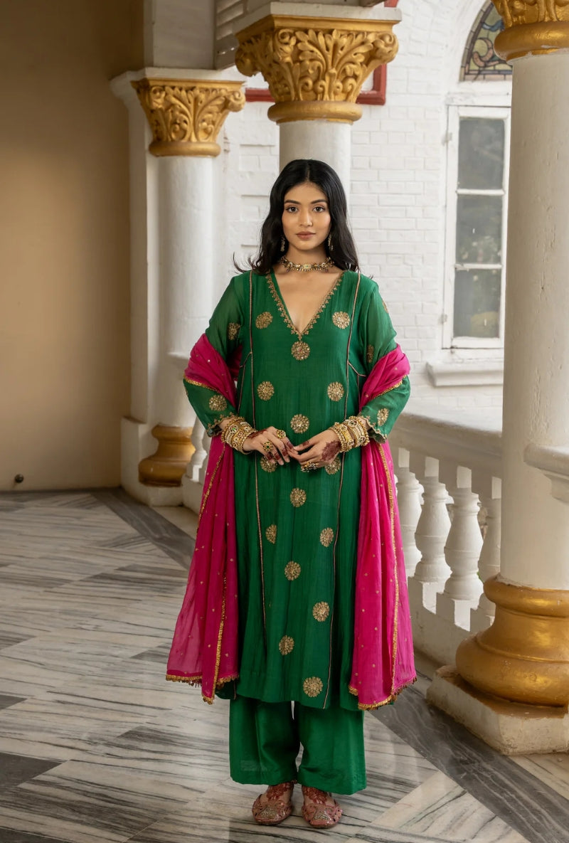 Woman in a green and pink traditional outfit standing in an elegant interior setting.
