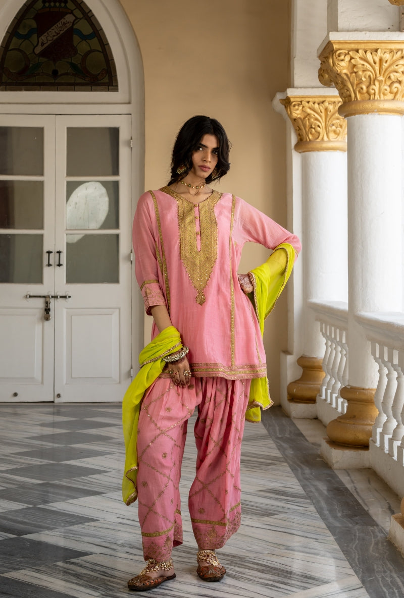 Woman in a pink traditional outfit with gold embroidery standing in a decorative interior setting.