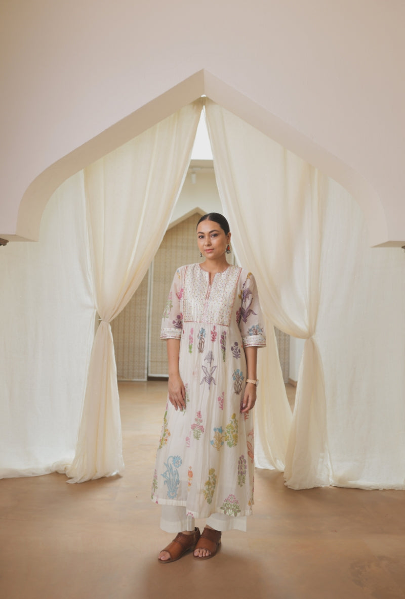 A woman stands in a doorway framed by draped cream fabric, wearing a long cream embroidered kurta with pastel multicolor floral prints and brown sandals.