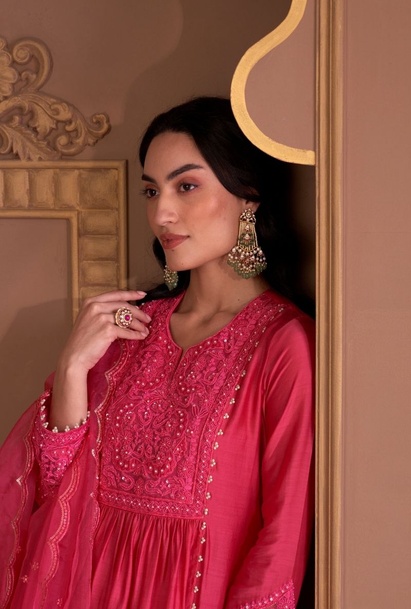 Woman wearing a pink embroidered traditional outfit with gold jewelry against a decorative wall.