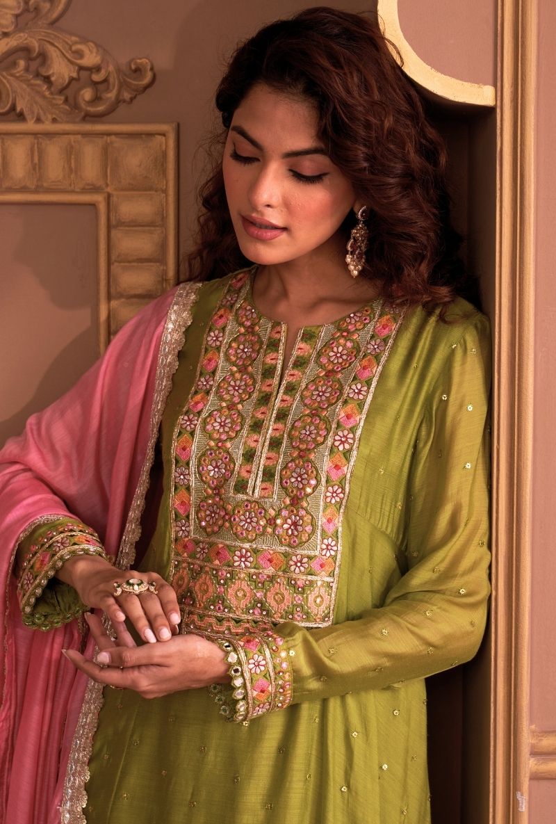 Woman in a green embroidered traditional outfit with a pink dupatta, standing indoors.