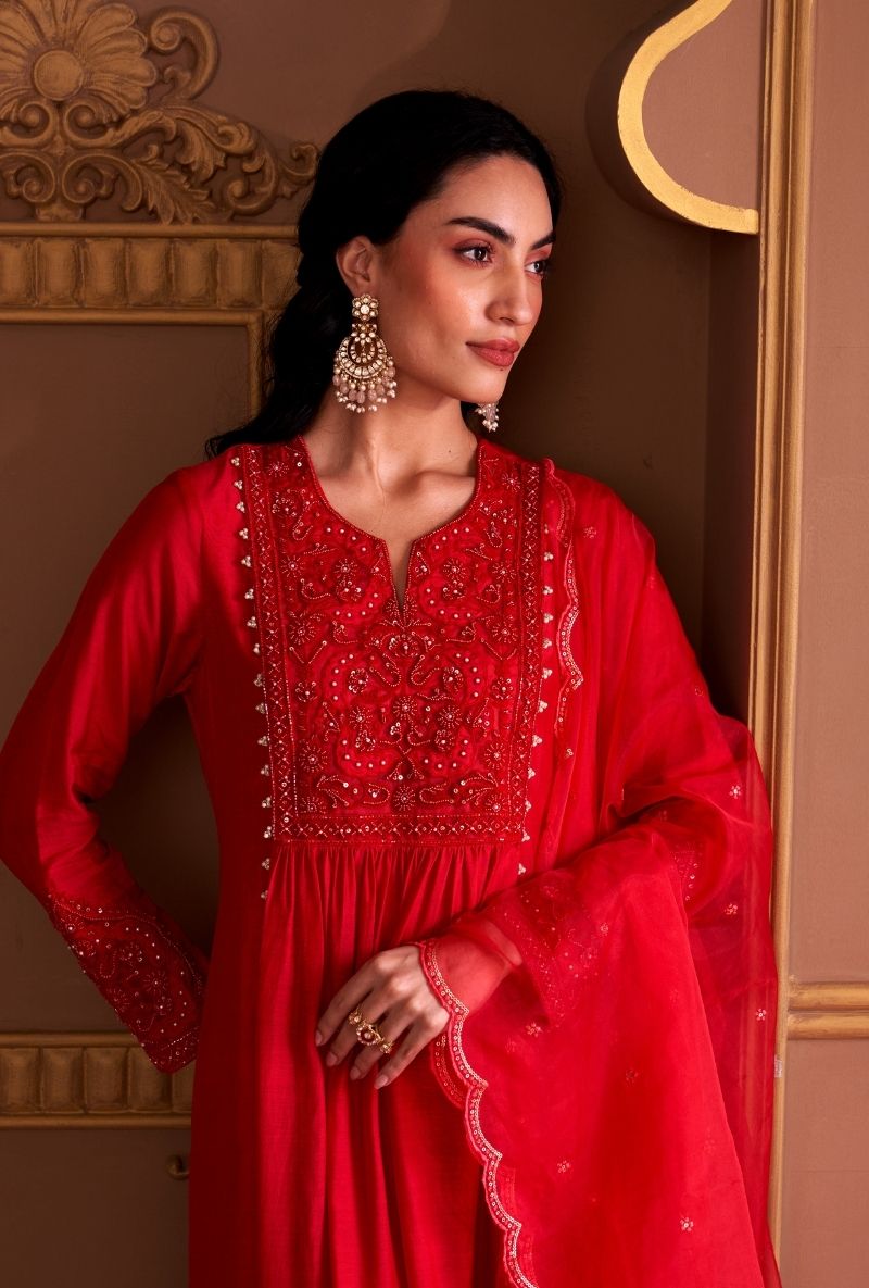 Woman in a red traditional outfit with intricate patterns and jewelry, standing in front of a decorative wall.