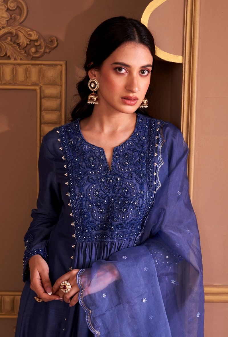 Woman wearing a blue embroidered outfit with jewelry against a decorative wall.