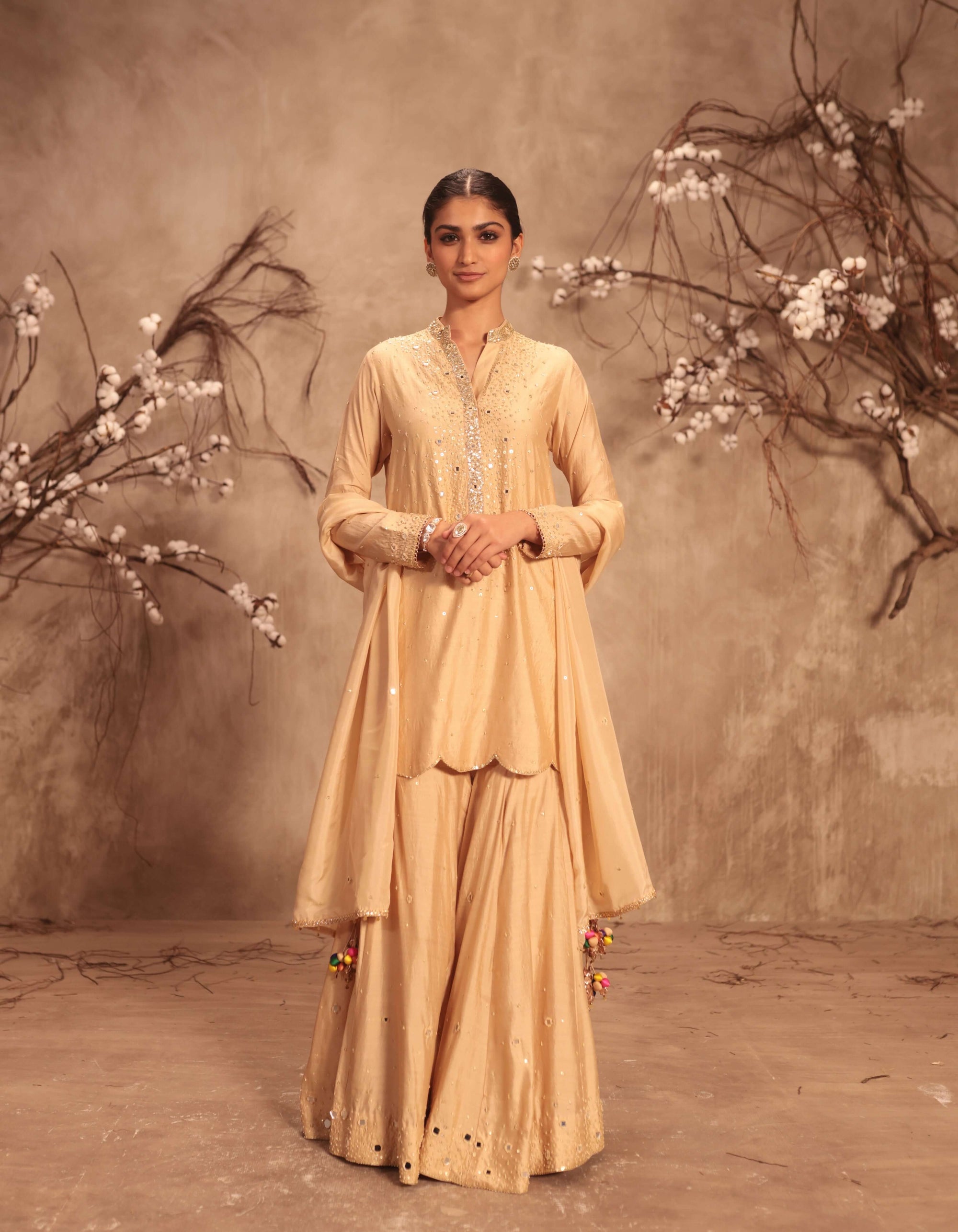 Woman in a beige traditional outfit standing against a textured wall with floral branches.