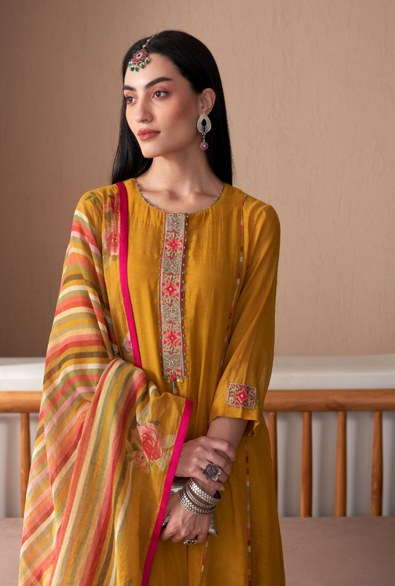 Woman wearing a mustard yellow traditional outfit with a colorful dupatta against a beige wall.